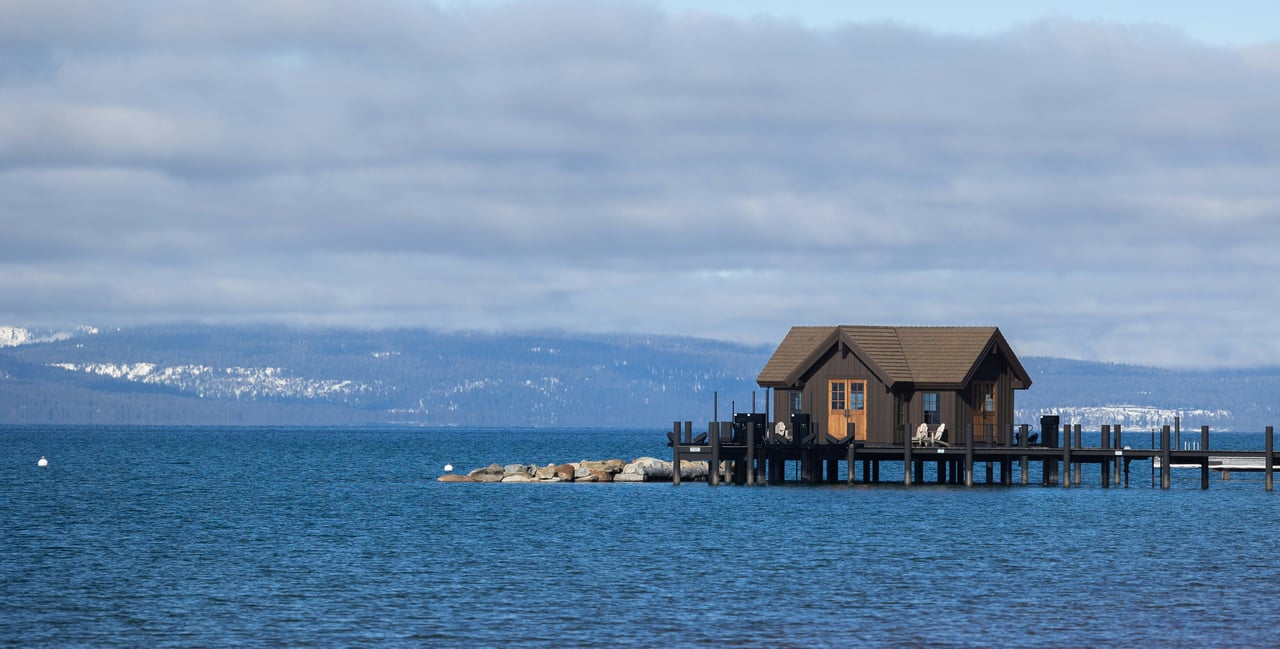 Dark-wood boathouse and dock over a lake, backed by snow-dusted mountains.