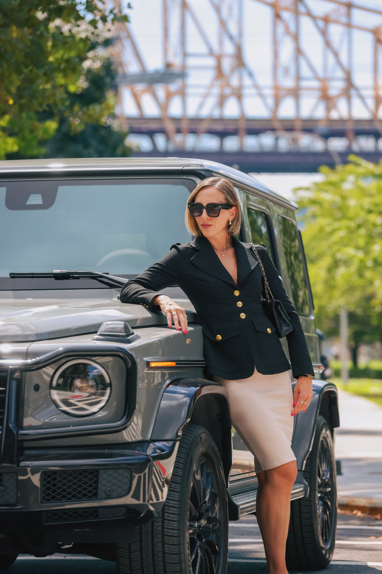 Woman in a black blazer and sunglasses leaning against a gray SUV beneath a steel bridge.