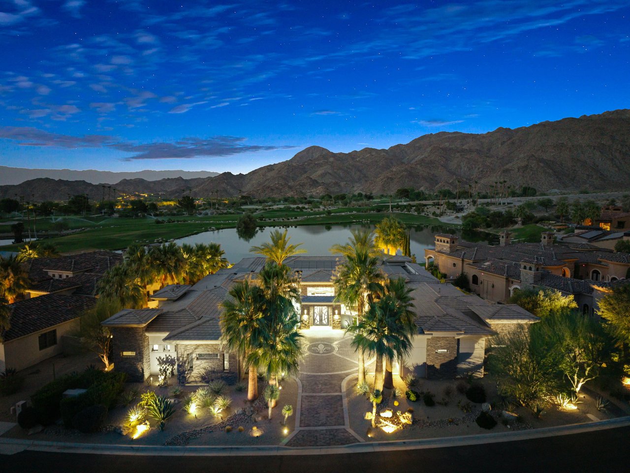 Aerial view of a desert home with illuminated palms, a lake, golf course, and mountains.