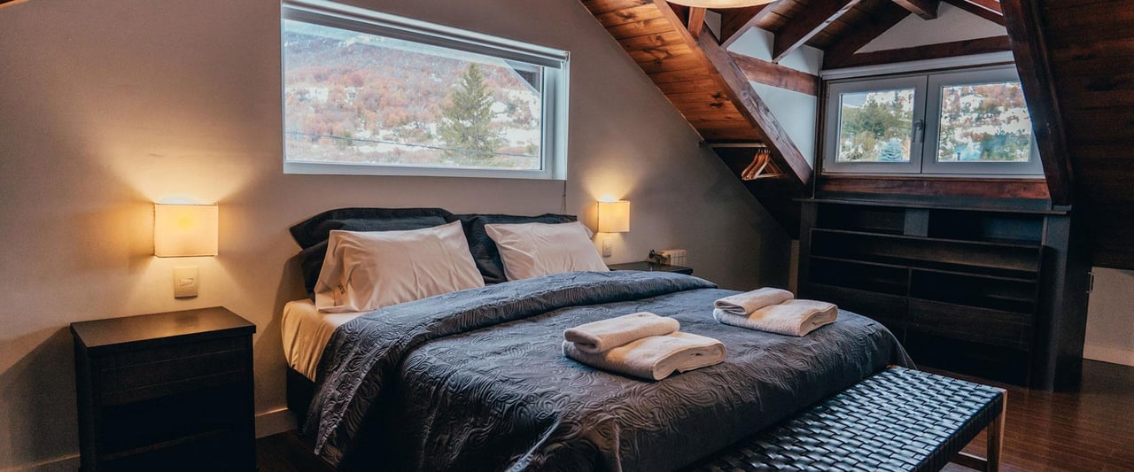 Attic bedroom with a large bed, folded towels, exposed timber beams, and windows overlooking trees.