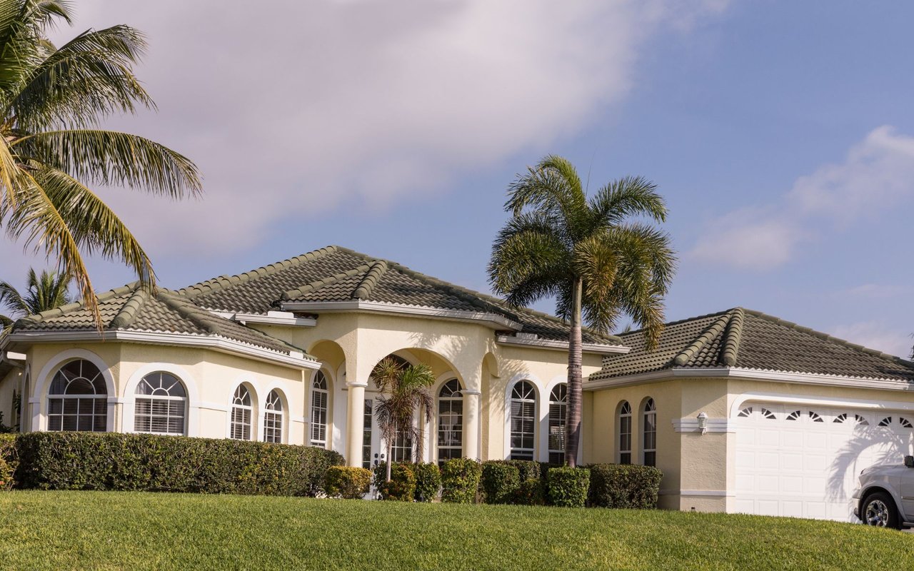 Cream stucco house with a tiled roof, arched windows, palm trees, and an attached garage.