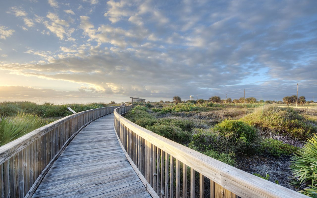 Wooden boardwalk winding through coastal vegetation beneath a dramatic sunrise sky.