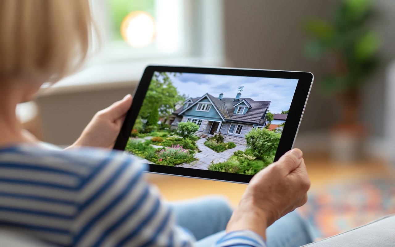 Person views a two-story house and landscaped garden on a tablet during a virtual showing.