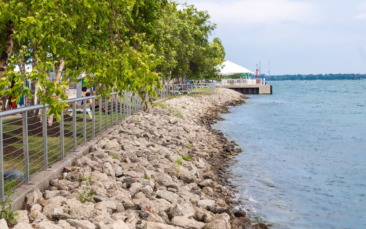Tree-lined waterfront promenade beside a rocky shoreline and pavilion.
