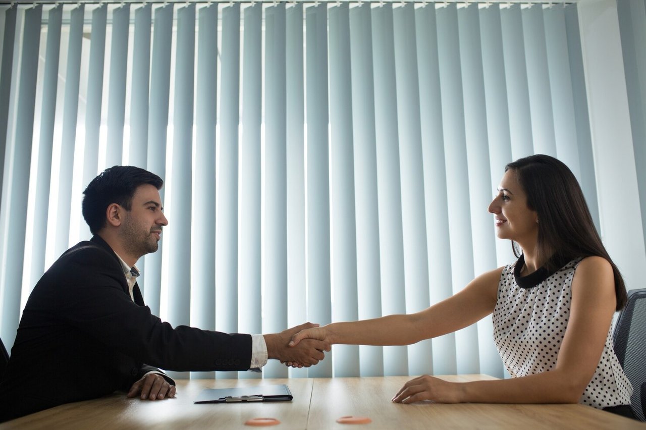 A man and woman shake hands across a table during a business meeting.