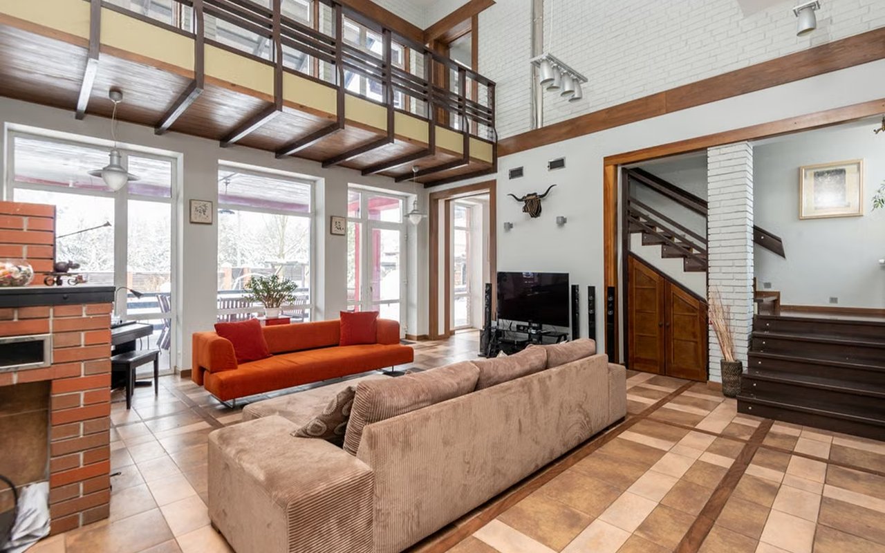 Double-height living room with a brick fireplace, mezzanine balcony, tiled floors, and colorful sofas.