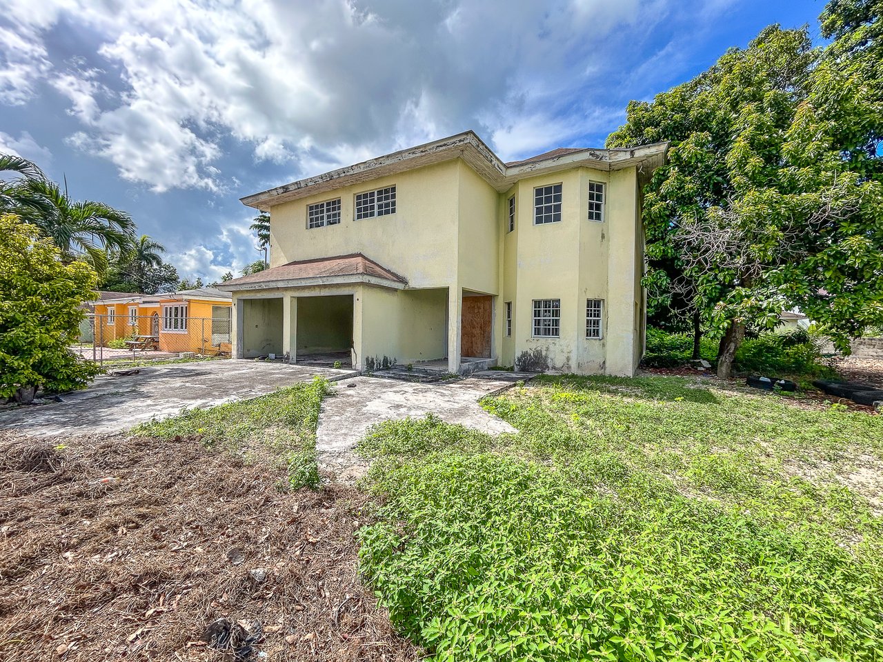 Two-story pale yellow house with a covered carport, concrete driveway, and overgrown lawn.