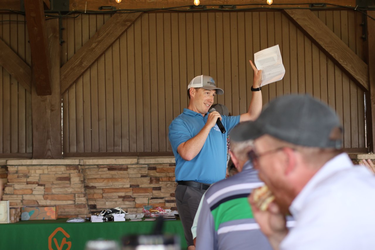 A man speaks into a microphone and raises a printed sheet before an audience.