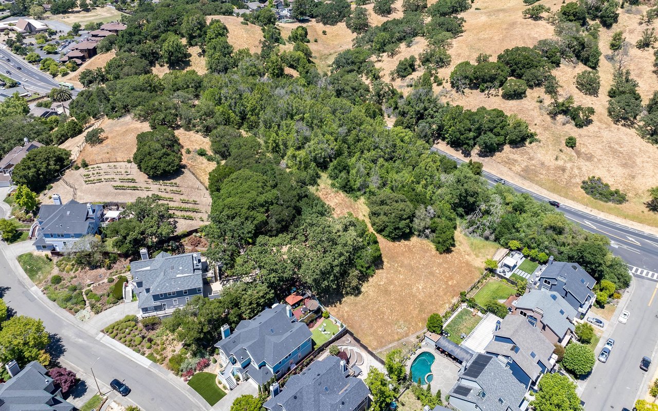 Aerial neighborhood with homes, tree-lined yards, a swimming pool, and dry open land.