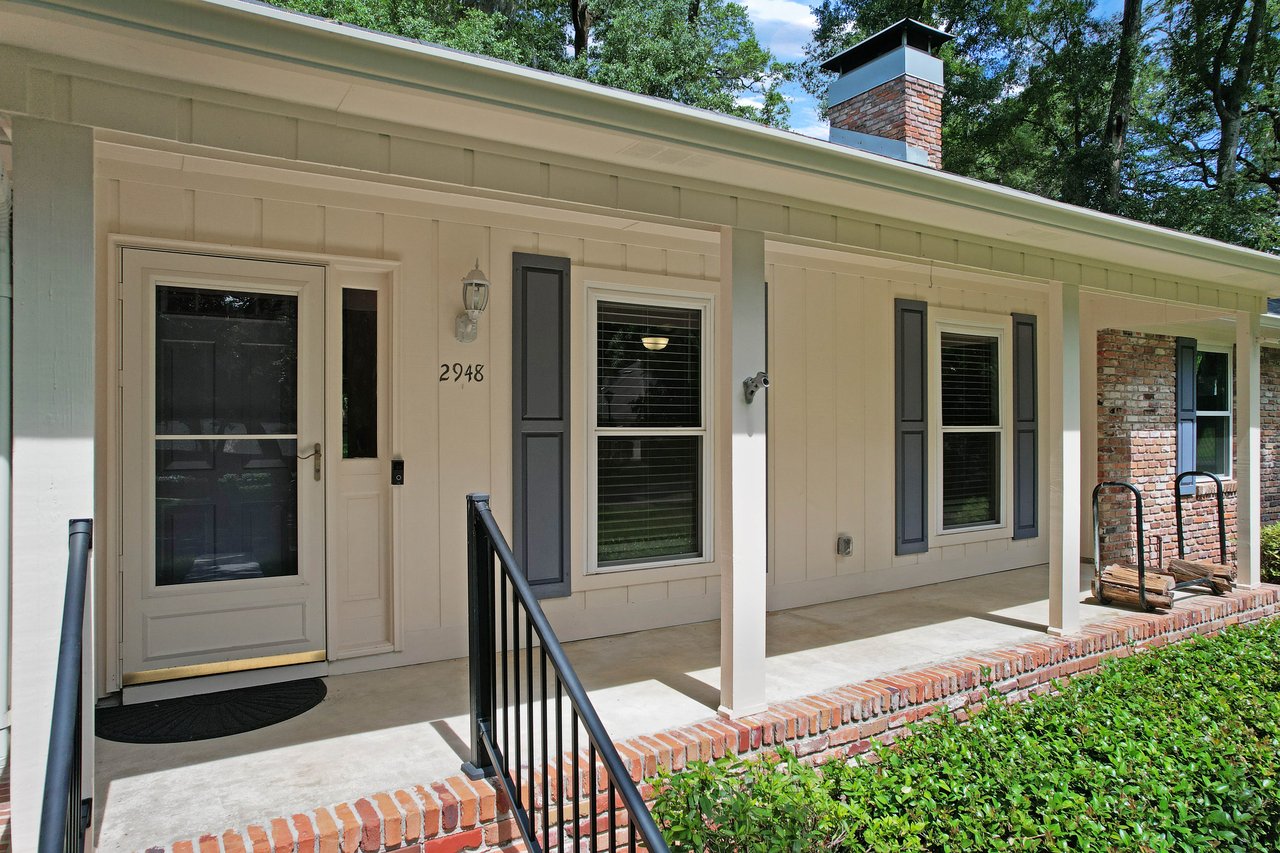 Front porch of a house with white siding, large windows with shutters, and a brick chimney. A step leads to the door, surrounded by greenery.