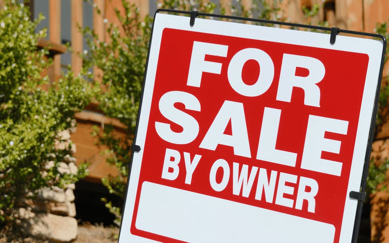 Red and white For Sale By Owner sign in front of a leafy garden.