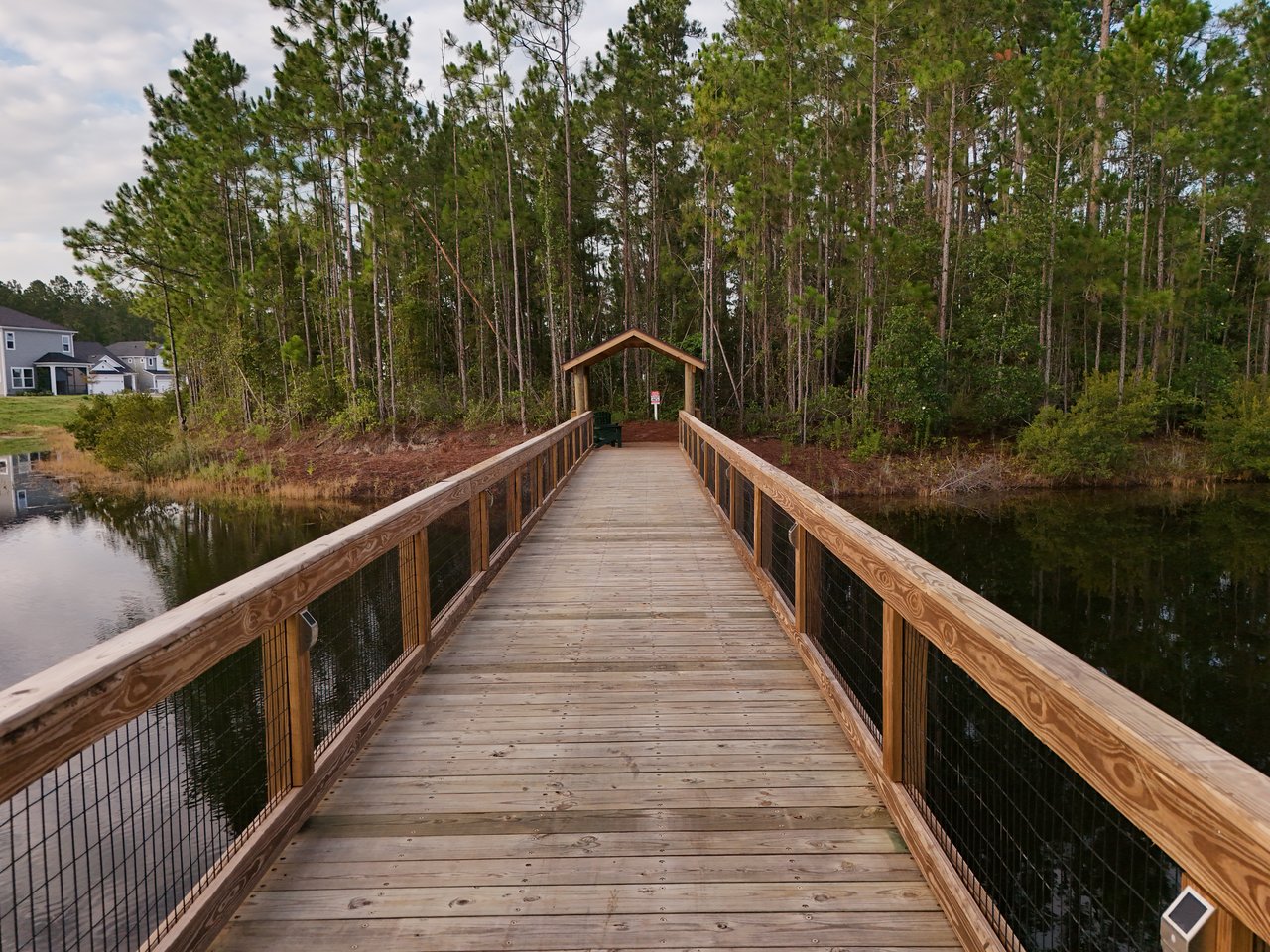 Wooden boardwalk crosses a pond toward a covered entrance beside a wooded area.