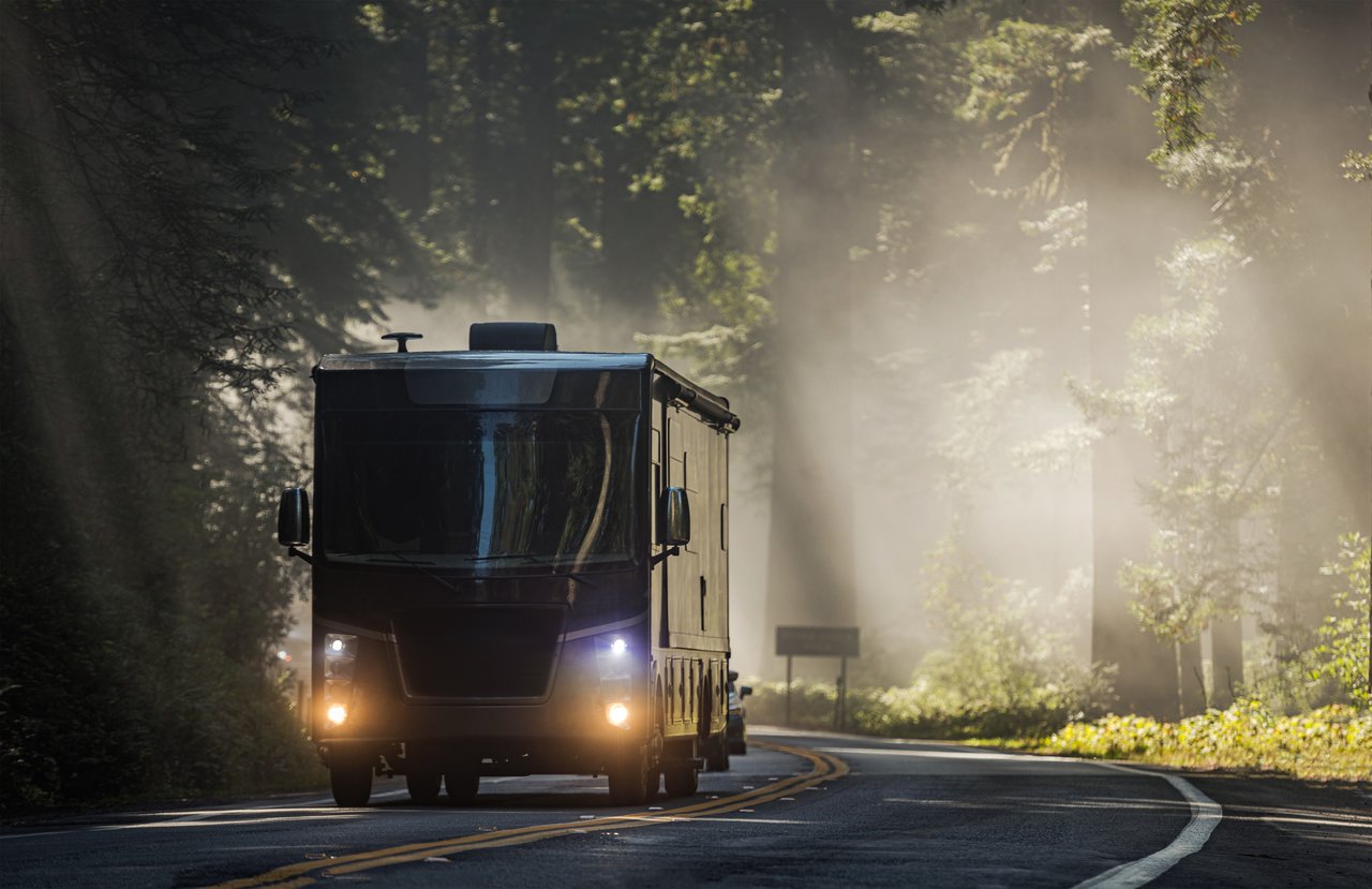 A large, modern luxury class A motorhome driving down a scenic, misty road winding through a dense forest.