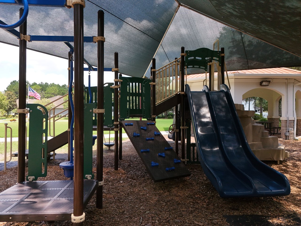 Community playground with a shaded climbing structure and double slide beside a pavilion.