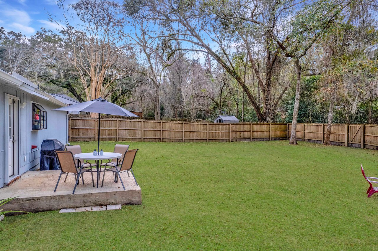 Spacious backyard with green lawn and tall trees. A patio set with an umbrella and chairs is on a concrete slab. Wooden fence encloses the area. Calm ambiance.