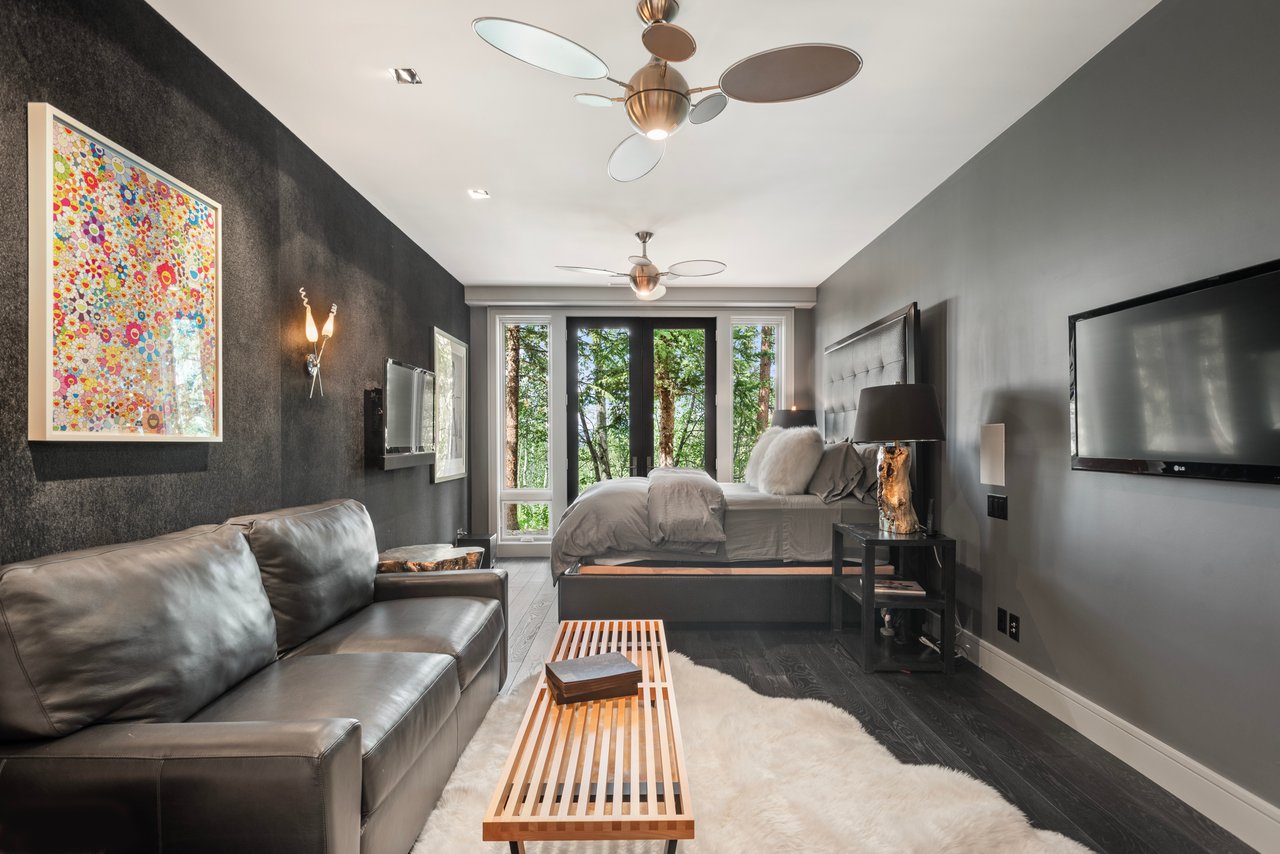 Bedroom with a leather sofa, colorful framed artwork, and windows overlooking leafy trees.