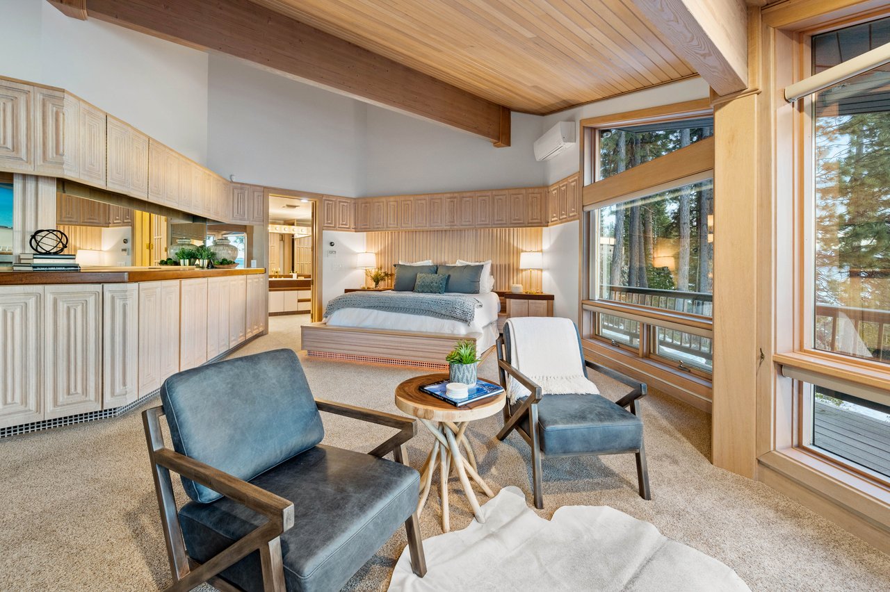 Bedroom suite with a vaulted wood ceiling, built-in cabinetry, lounge seating, and large windows overlooking trees.
