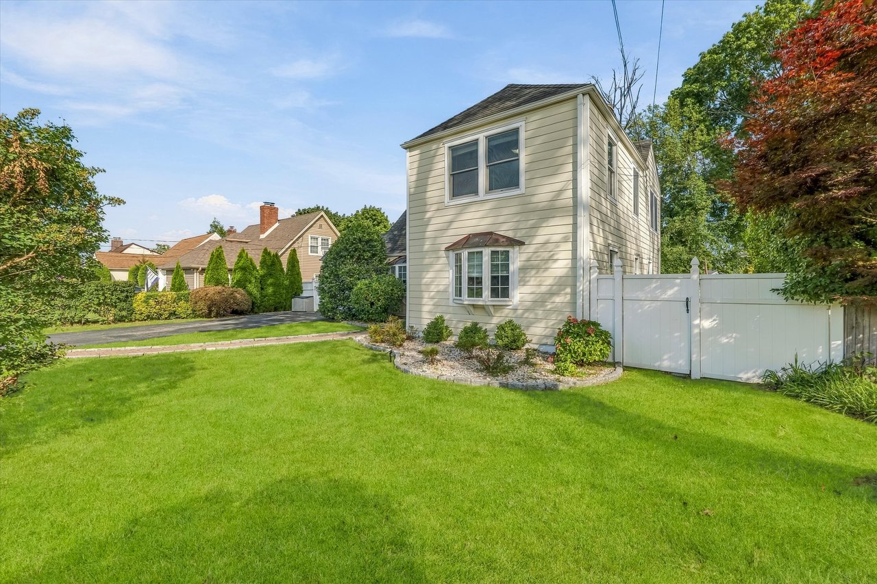 Side of a two-story house with a bay window, white fence, and broad green lawn.