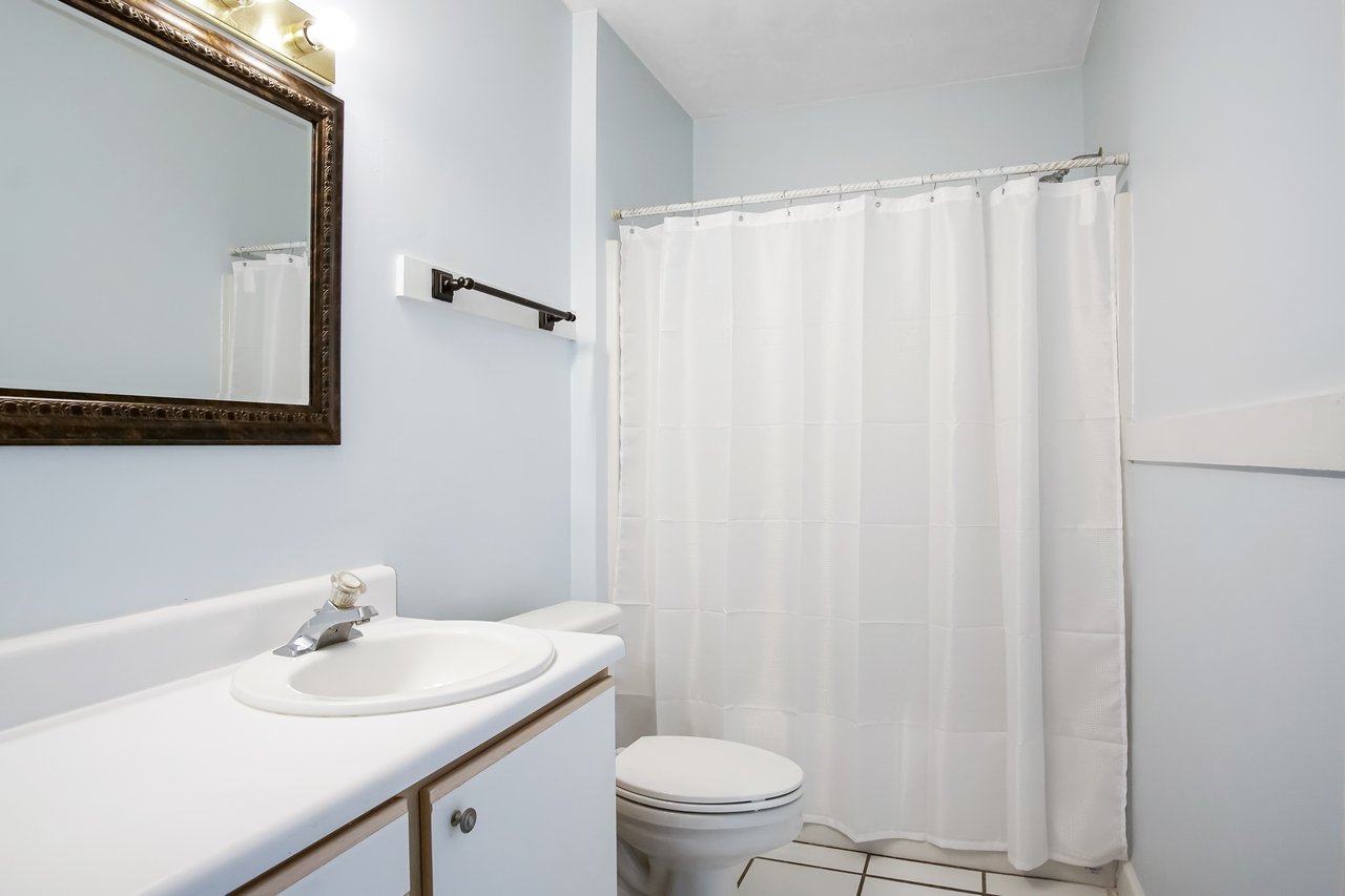 A clean, simple bathroom with a white countertop sink, modern faucet, and a large mirror. A toilet and white shower curtain are visible, conveying a minimalist mood.