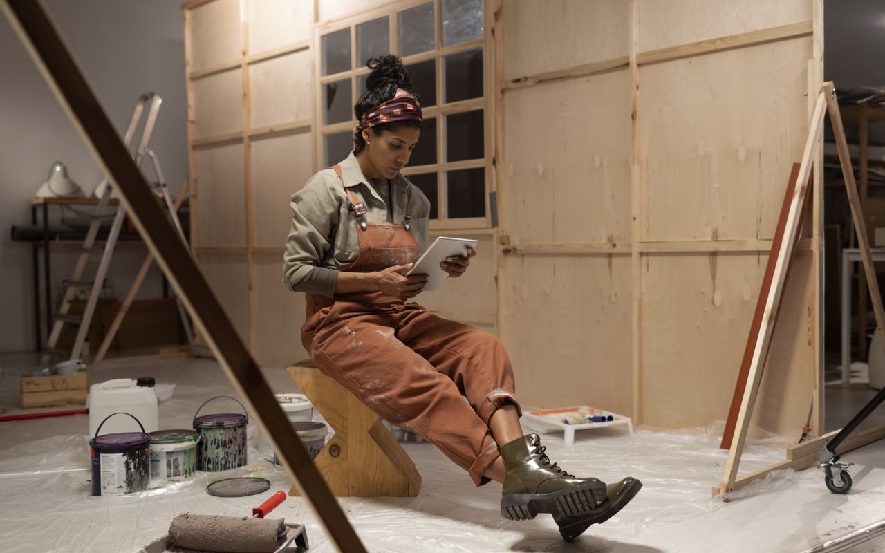 A woman in work clothes checks a tablet while seated in a room under renovation.