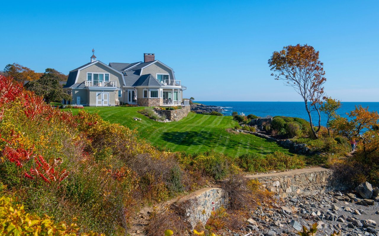 Coastal shingle home on a manicured lawn above a rocky ocean shoreline.