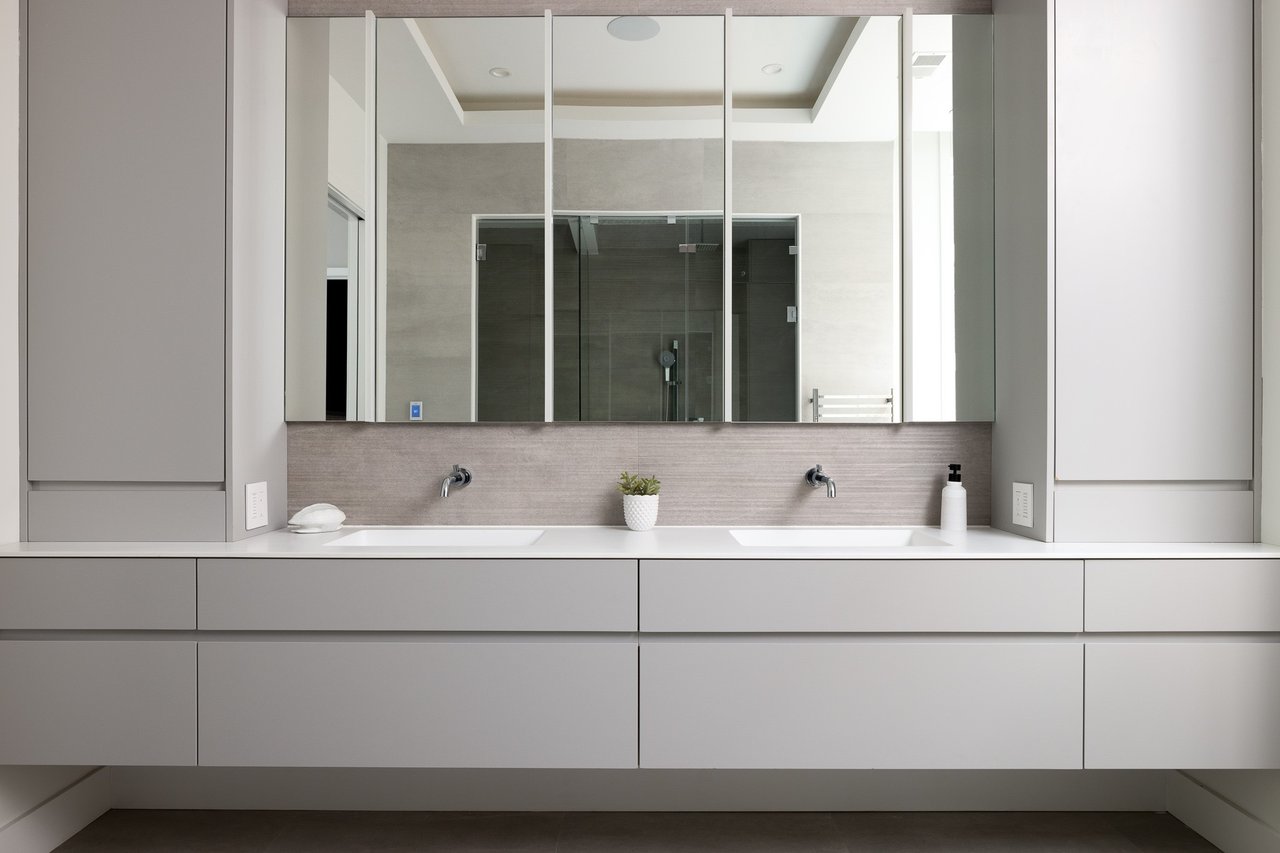 Modern double-sink bathroom vanity with gray cabinetry, wall-mounted faucets, and a mirrored cabinet reflecting the shower.