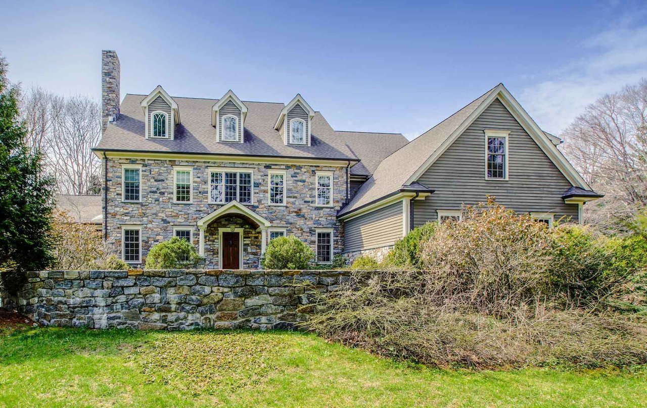 Stone-fronted house with three dormers, a gray gabled wing, and a low stone wall.