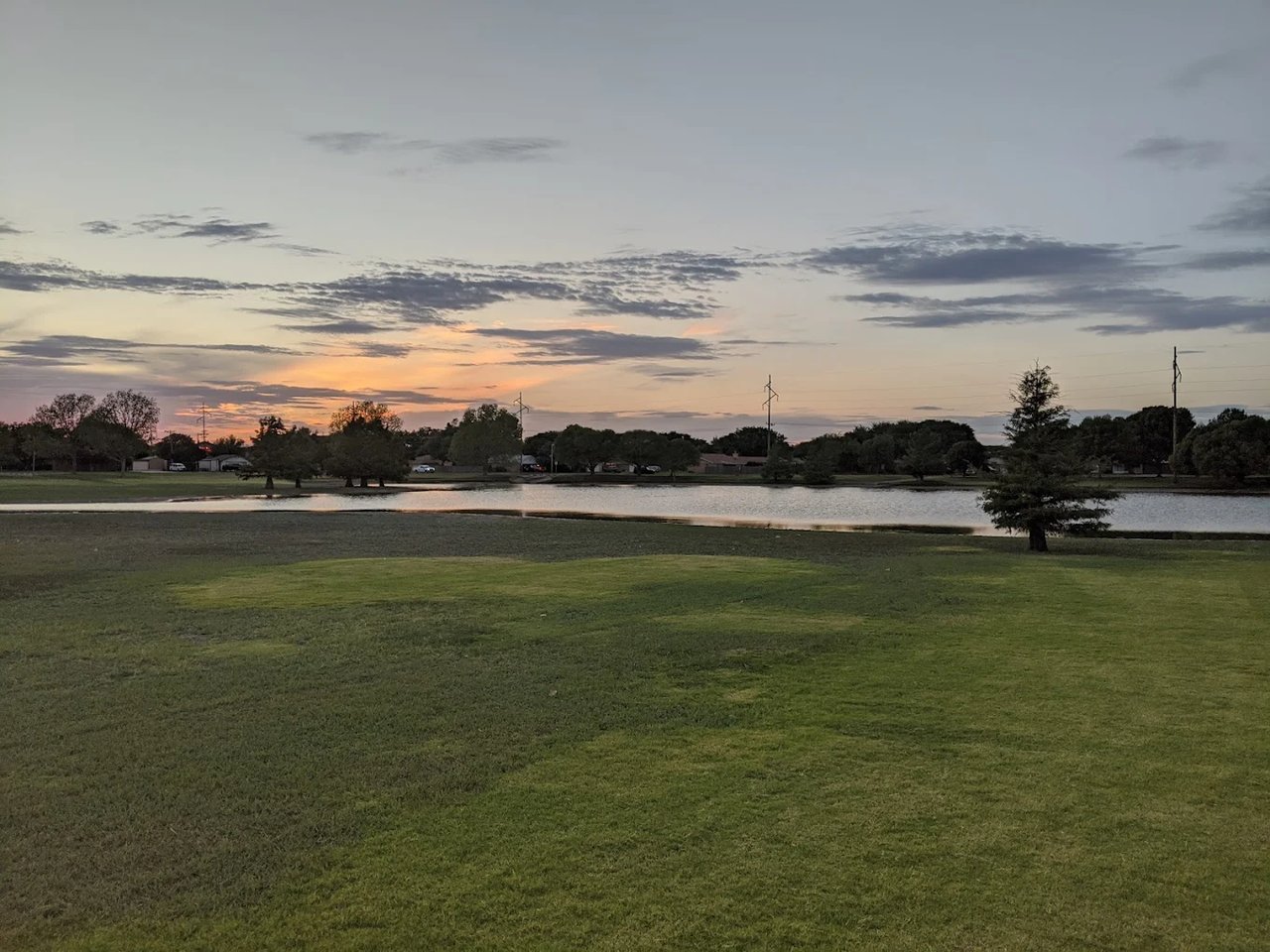 Neighborhood pond bordered by grassy parkland and trees beneath a colorful dusk sky.