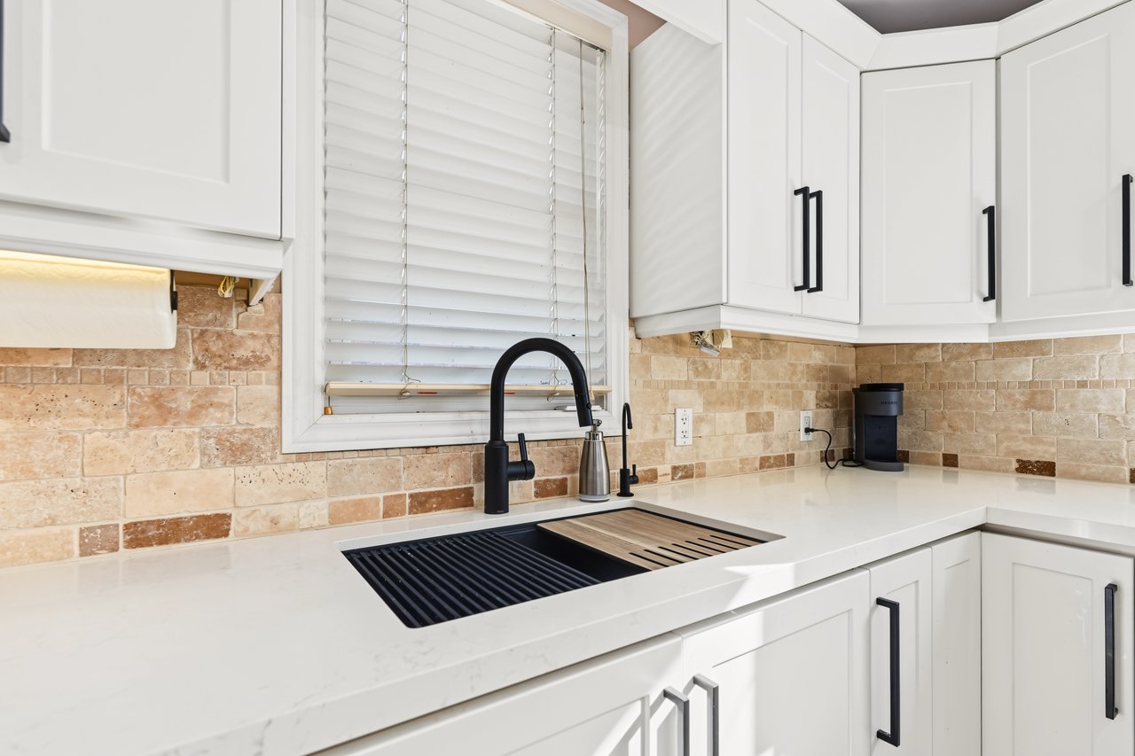 Kitchen with white cabinets, stone-tile backsplash, and black sink beneath a window with blinds.