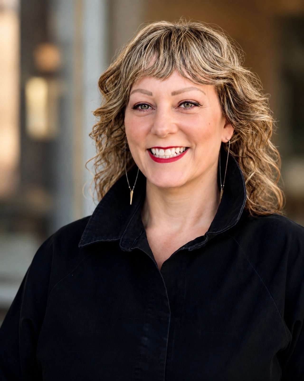 Jen Cowen smiling in a black coat, with curly light-brown hair and long drop earrings.