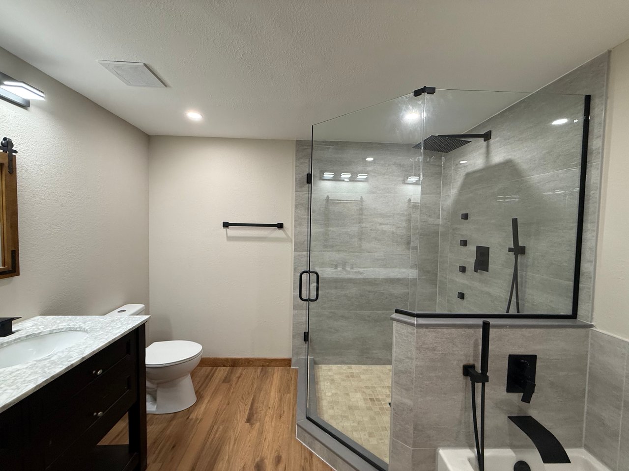 Bathroom with a glass-enclosed gray-tile shower, matte black fixtures, dark vanity, and wood-look flooring.