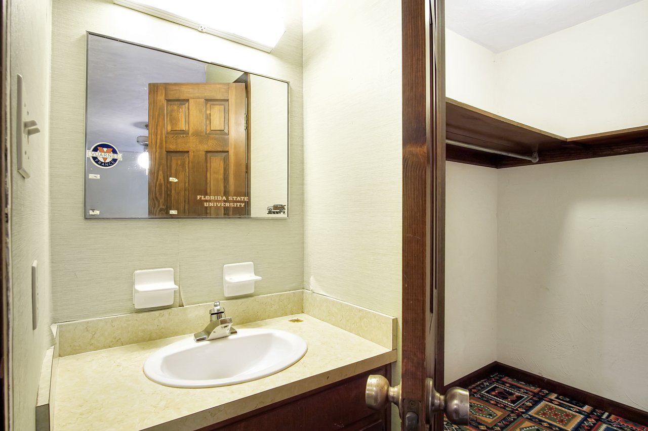 Small bathroom with beige walls featuring a mirror reflecting a wooden door. A white sink is set in a cream countertop. An open closet is visible beside the vanity.