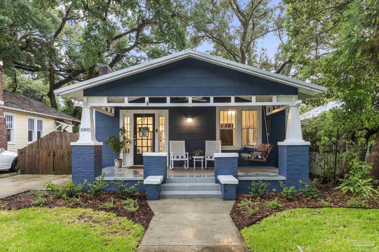 Blue Craftsman bungalow with a deep covered porch, white columns, and a porch swing beneath mature trees.