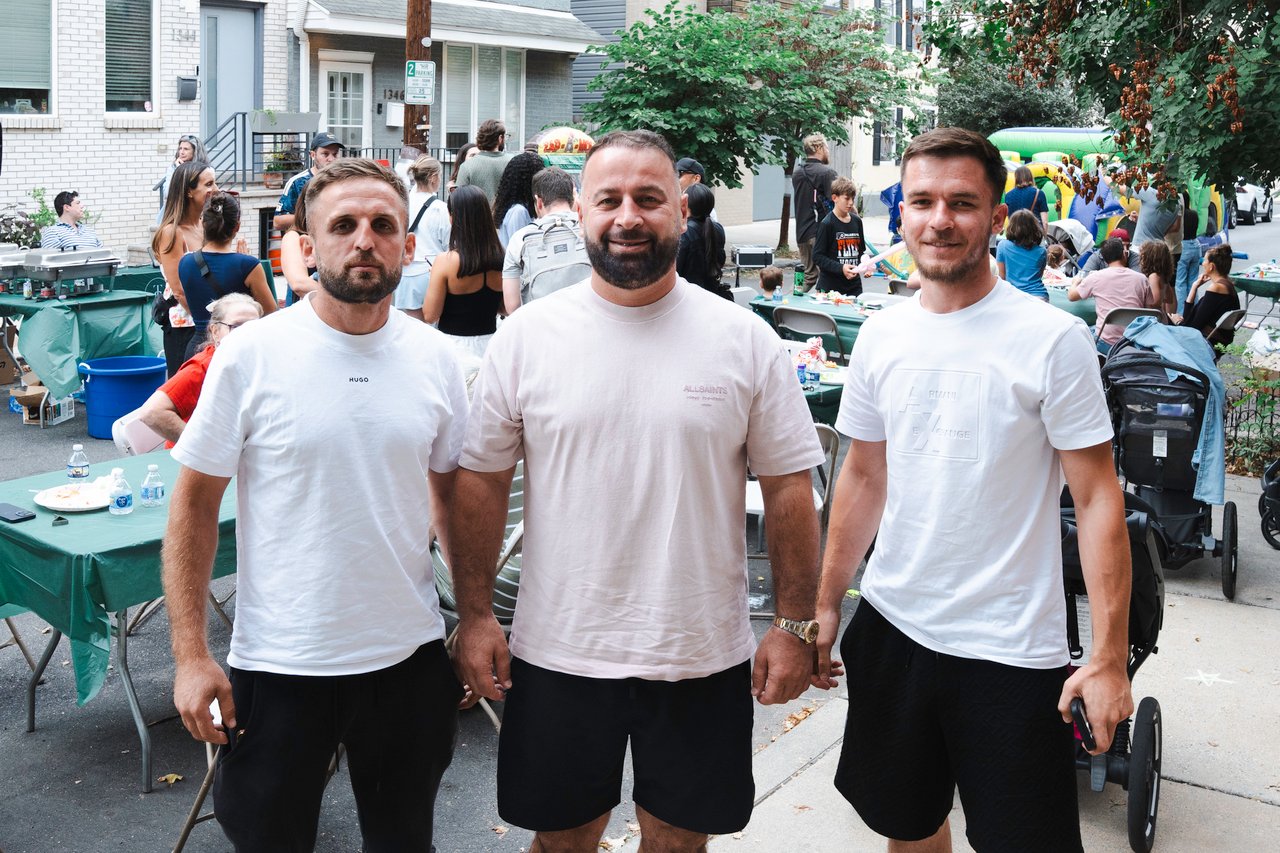 Three men pose together at a neighborhood block party, with guests and an inflatable play area behind them.