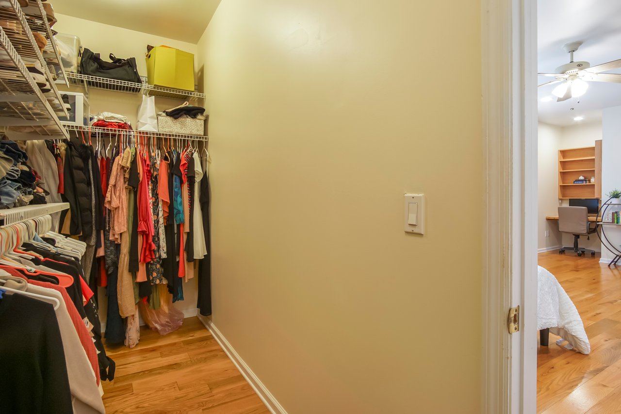 A walk-in closet with neatly hung clothes on racks. The closet opens into a softly lit hallway, revealing a glimpse of a cozy, furnished room.