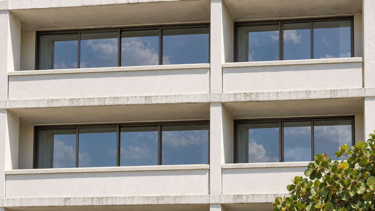 Close upward view of an off-white condominium facade with recessed balconies, bronze-framed glass doors, and a sea grape crown.
