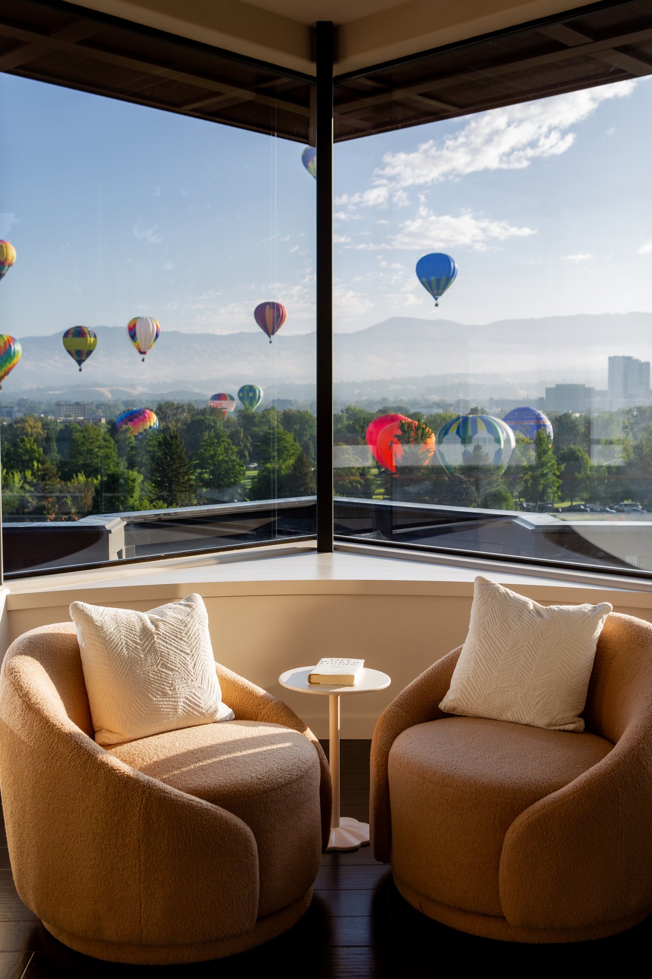 Two tan chairs sit beside corner windows overlooking hot air balloons, the city, and distant mountains.