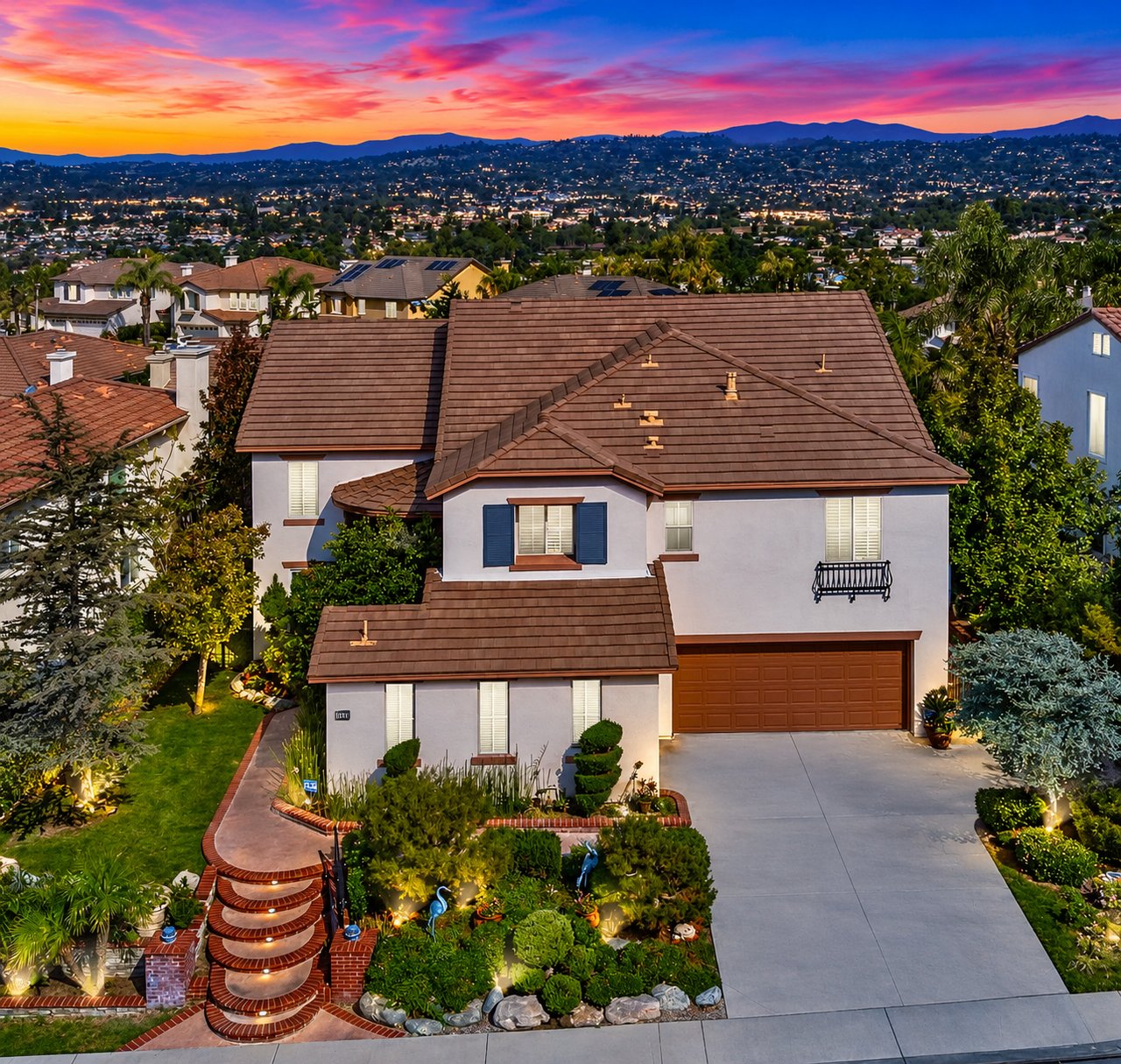 Two-story stucco home with a tile roof, landscaped yard, and curved brick steps at sunset.