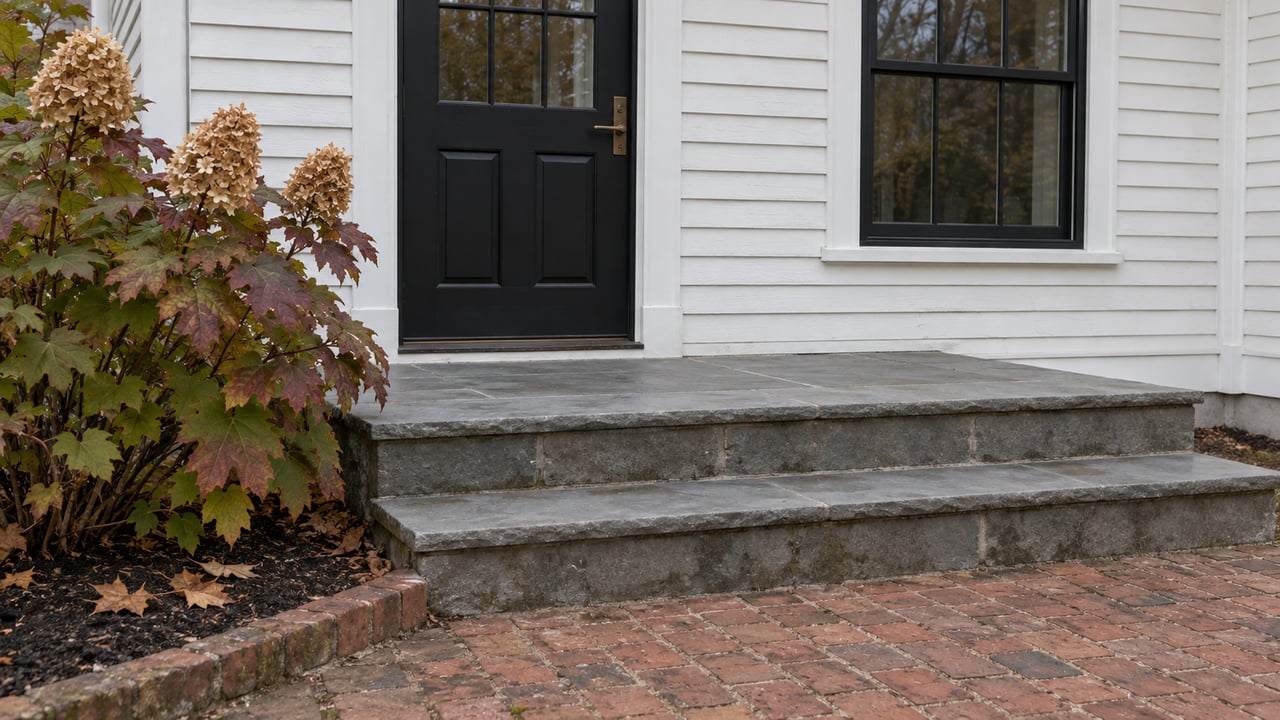 White clapboard entry with a black door, gray stone steps, brick walk, and an oakleaf hydrangea turning burgundy.