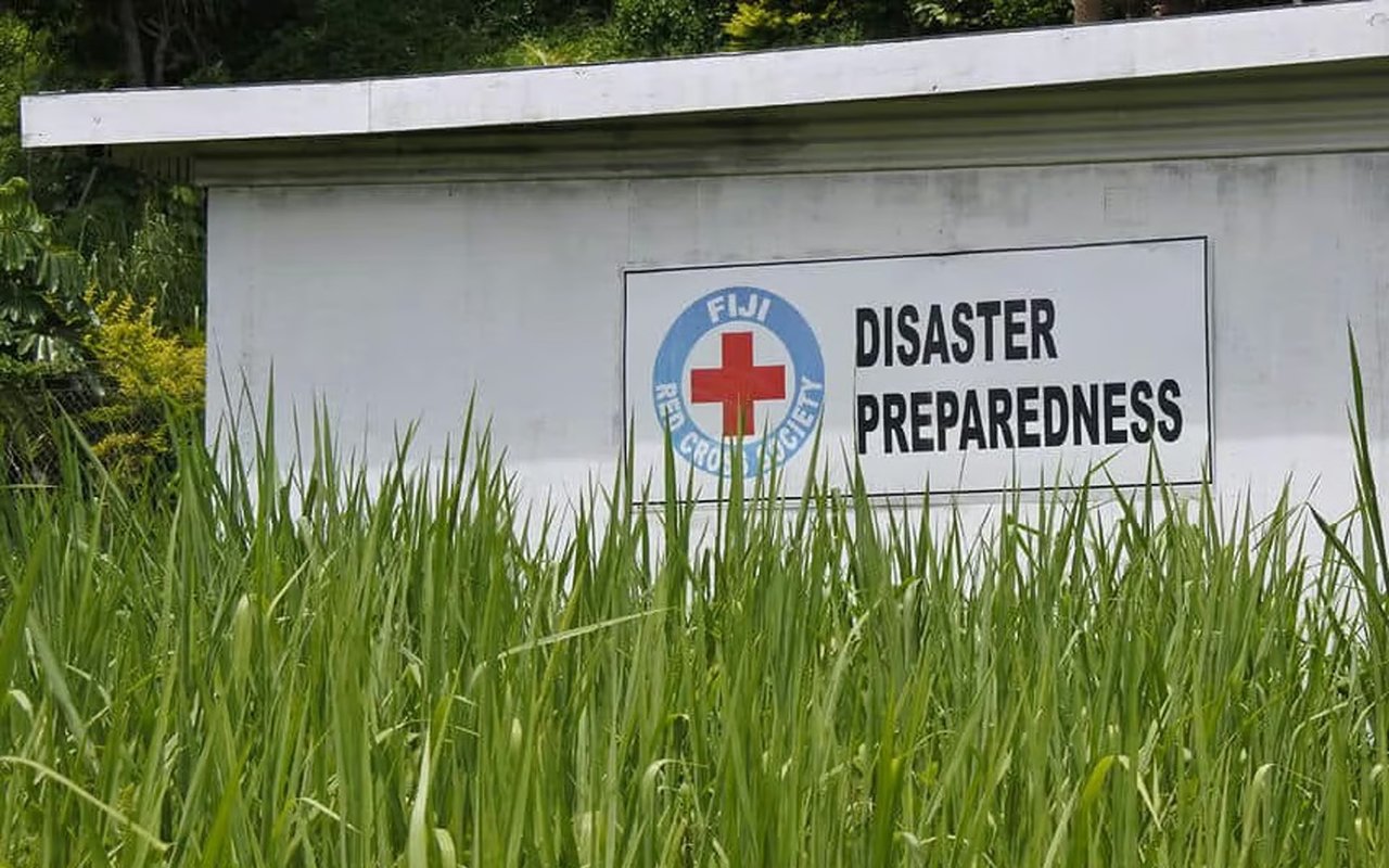 Disaster preparedness sign with the Fiji Red Cross emblem, partly hidden by tall grass.