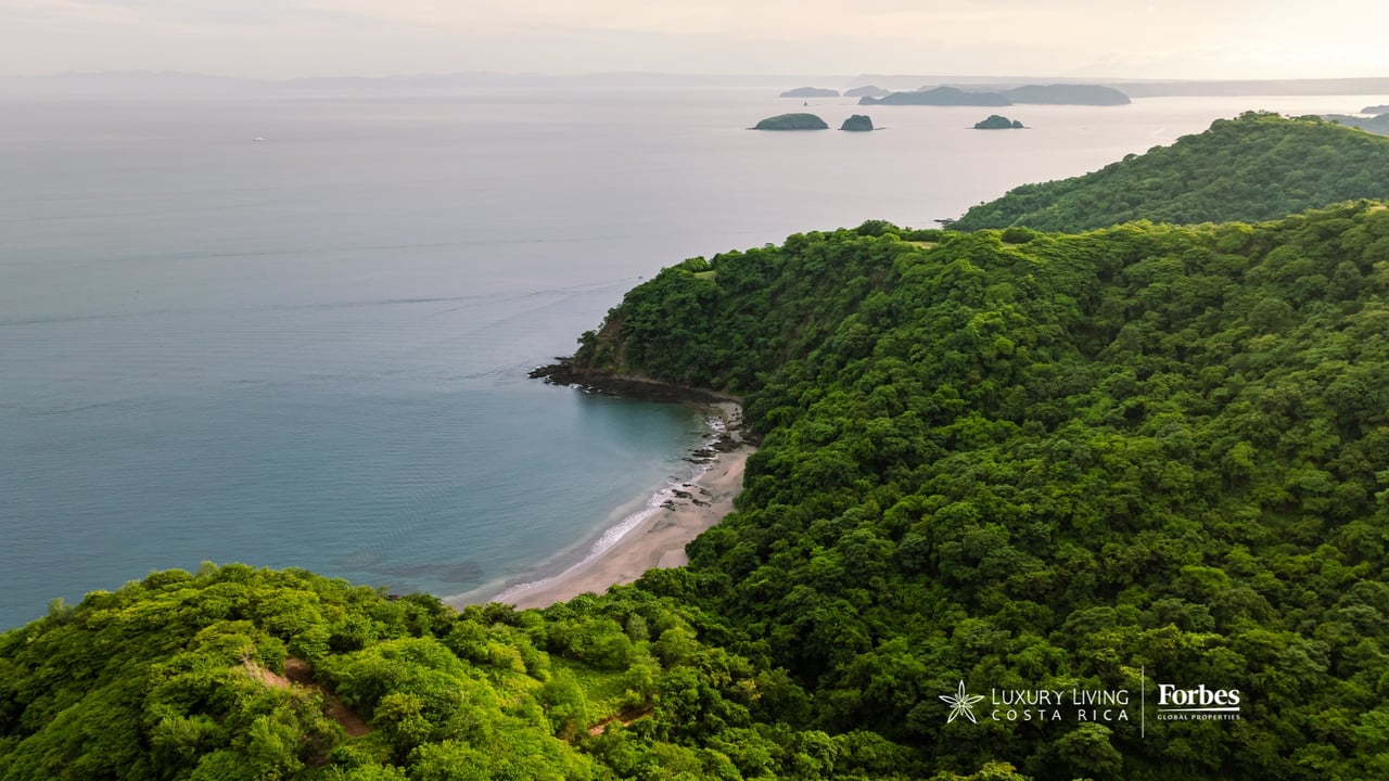 Aerial view of forested coastline, sandy cove, calm ocean, and offshore islands.