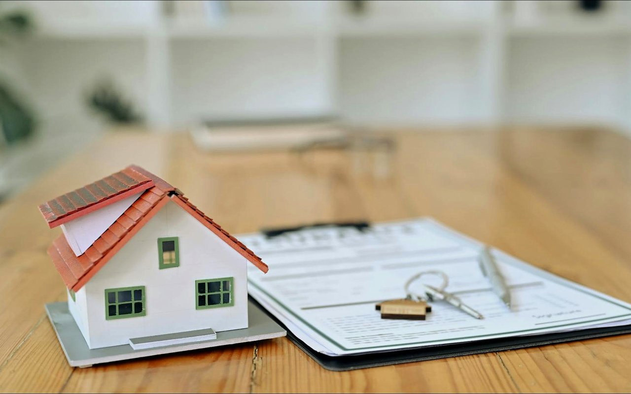 Miniature house beside mortgage paperwork and a key on a wooden table.