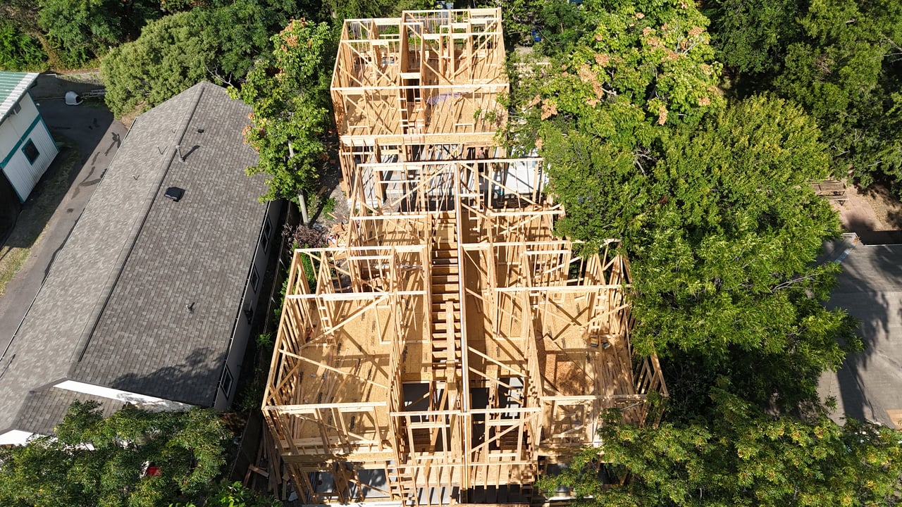 Wood-framed house under construction, viewed from above among mature trees and neighboring homes.