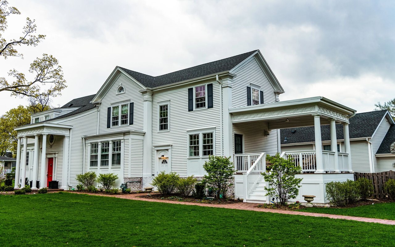 White two-story house with a red front door, covered porch, and broad green lawn.