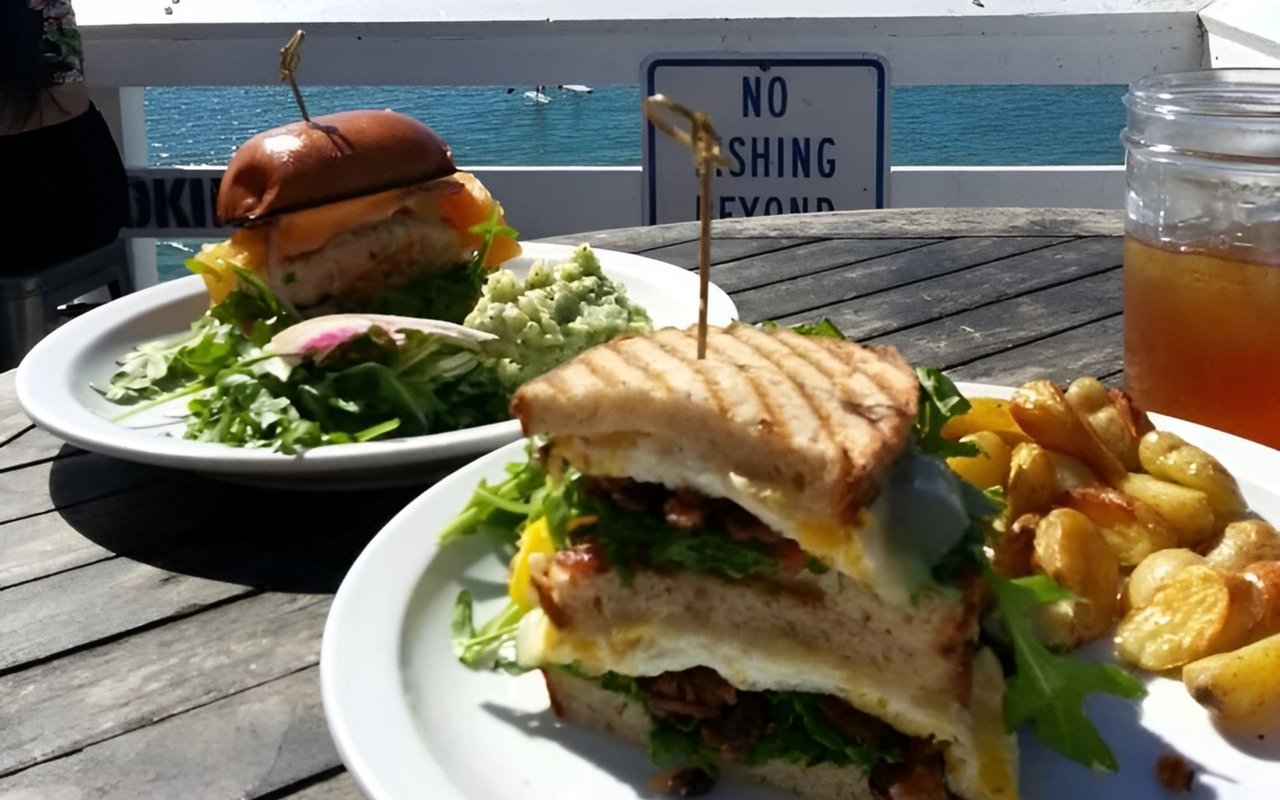 Two sandwiches with greens and roasted potatoes on an oceanfront outdoor table.