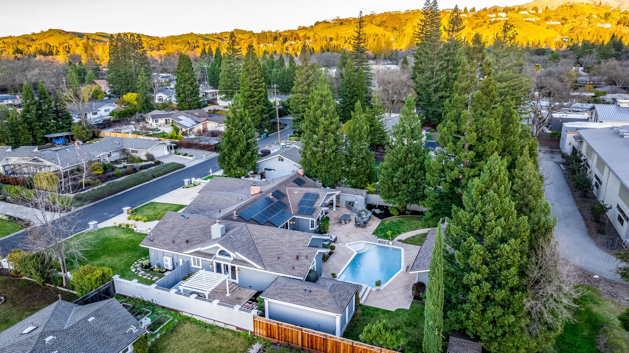 Aerial view of a tree-lined neighborhood, with a pool and solar panels in the foreground and golden hills beyond.