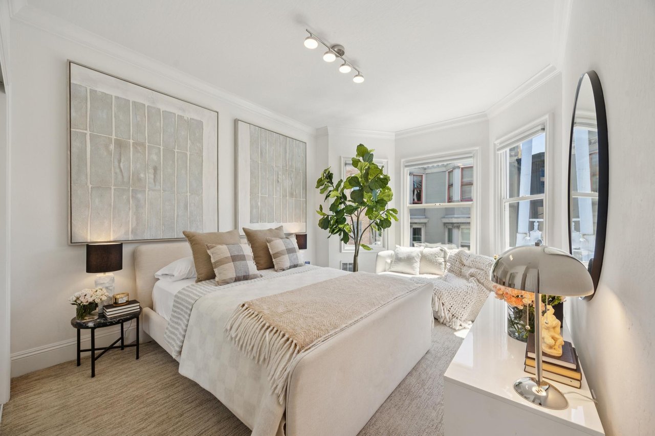 Staged bedroom with neutral bedding, large framed artwork, a potted tree, and tall windows.