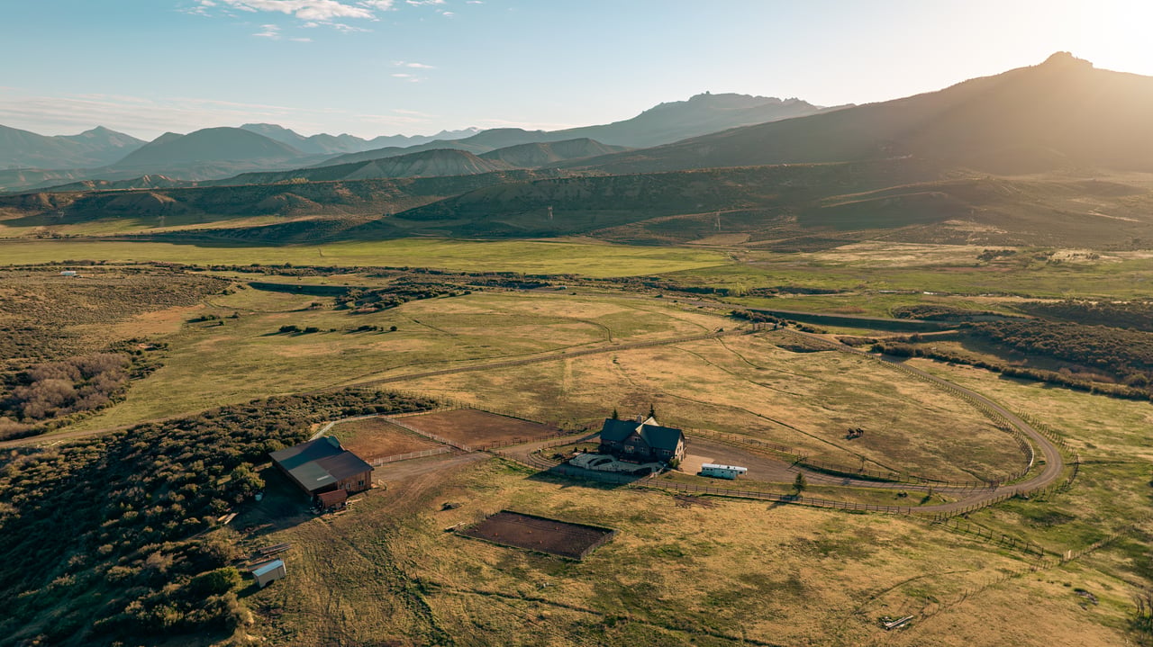 Aerial view of a ranch with a house, barns, fenced paddocks, open pasture, and mountains.