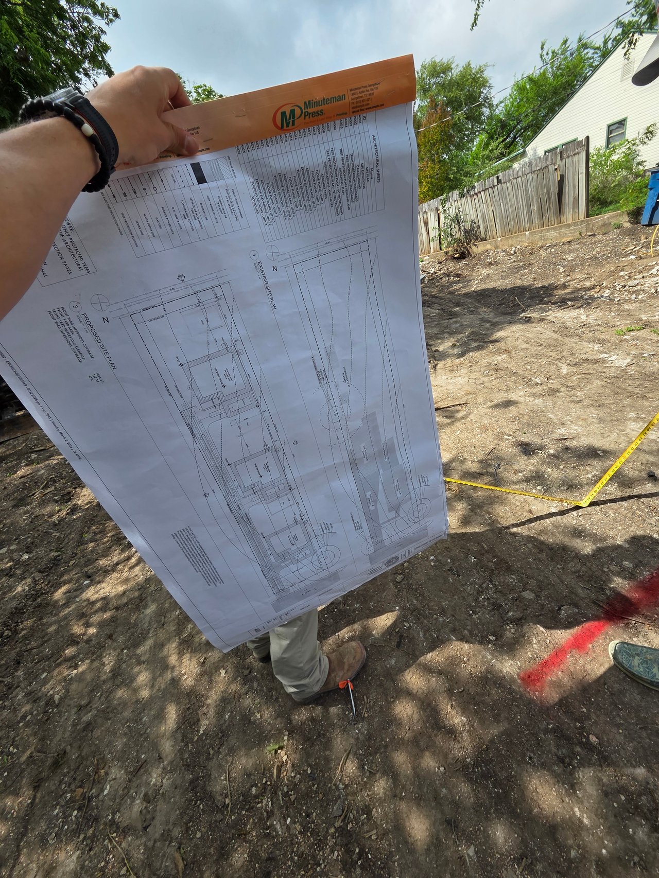 Residential development site with architectural plans held above a cleared lot marked with measuring tape.