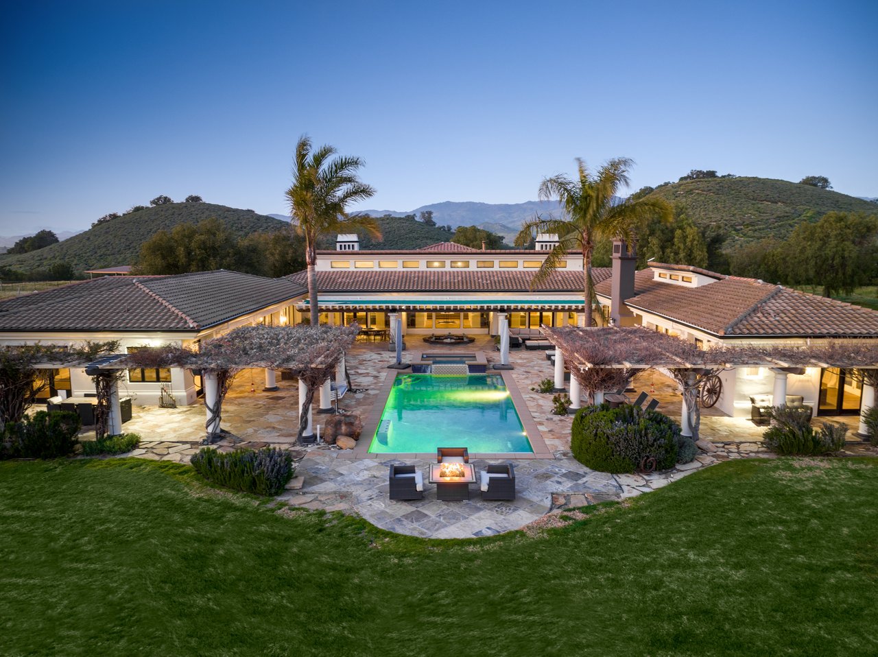Mediterranean-style home with illuminated pool and stone patio at dusk, backed by rolling hills.